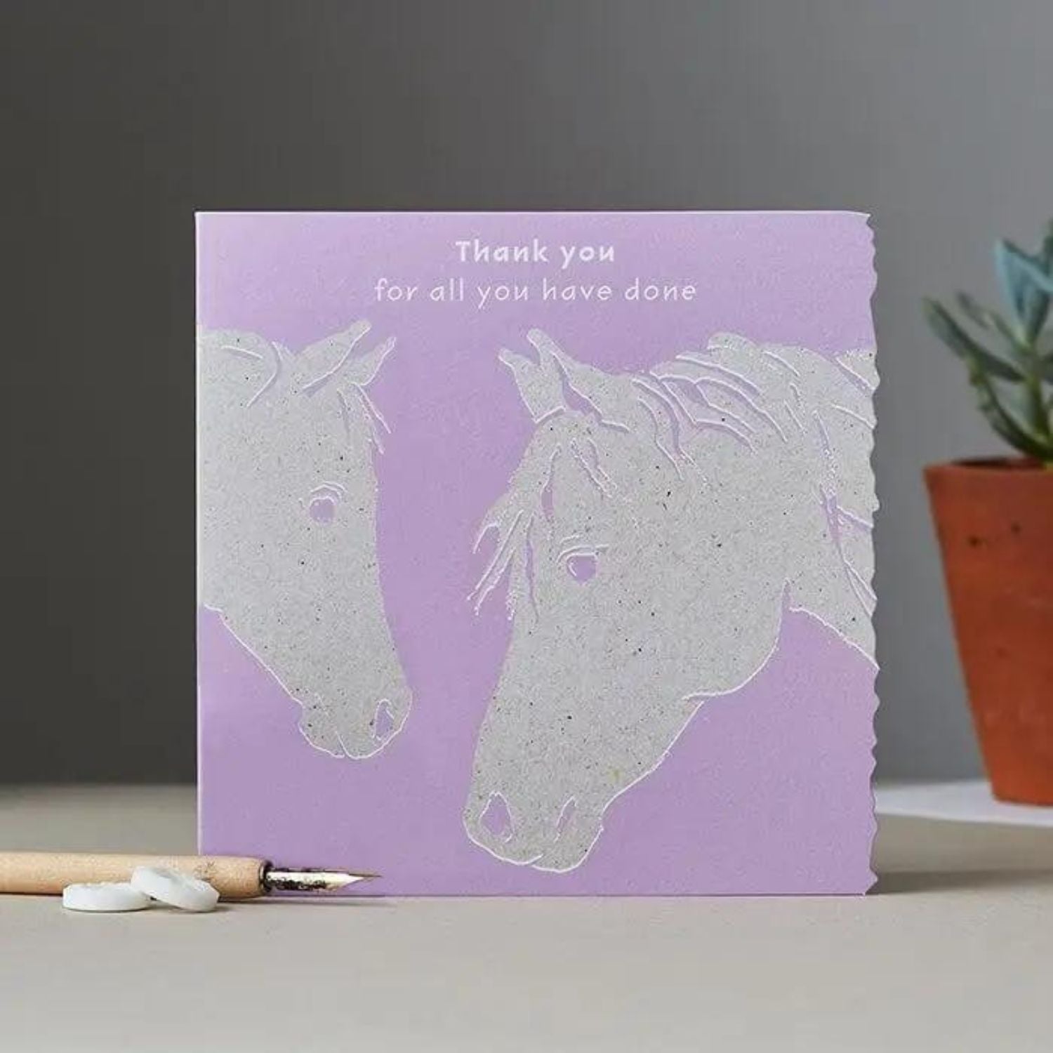 A purple card with two white horse illustrations facing each other and the text Thank you for all you have done above them. A pencil and plant are in the background.