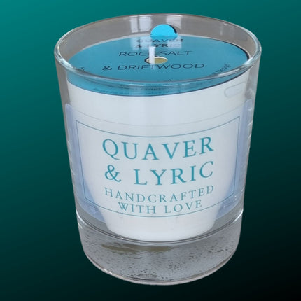 a white scented candle in a glass container with Quaver&Lyric Handcrafted with love label in teal on a transparent background. With a blue dust cover