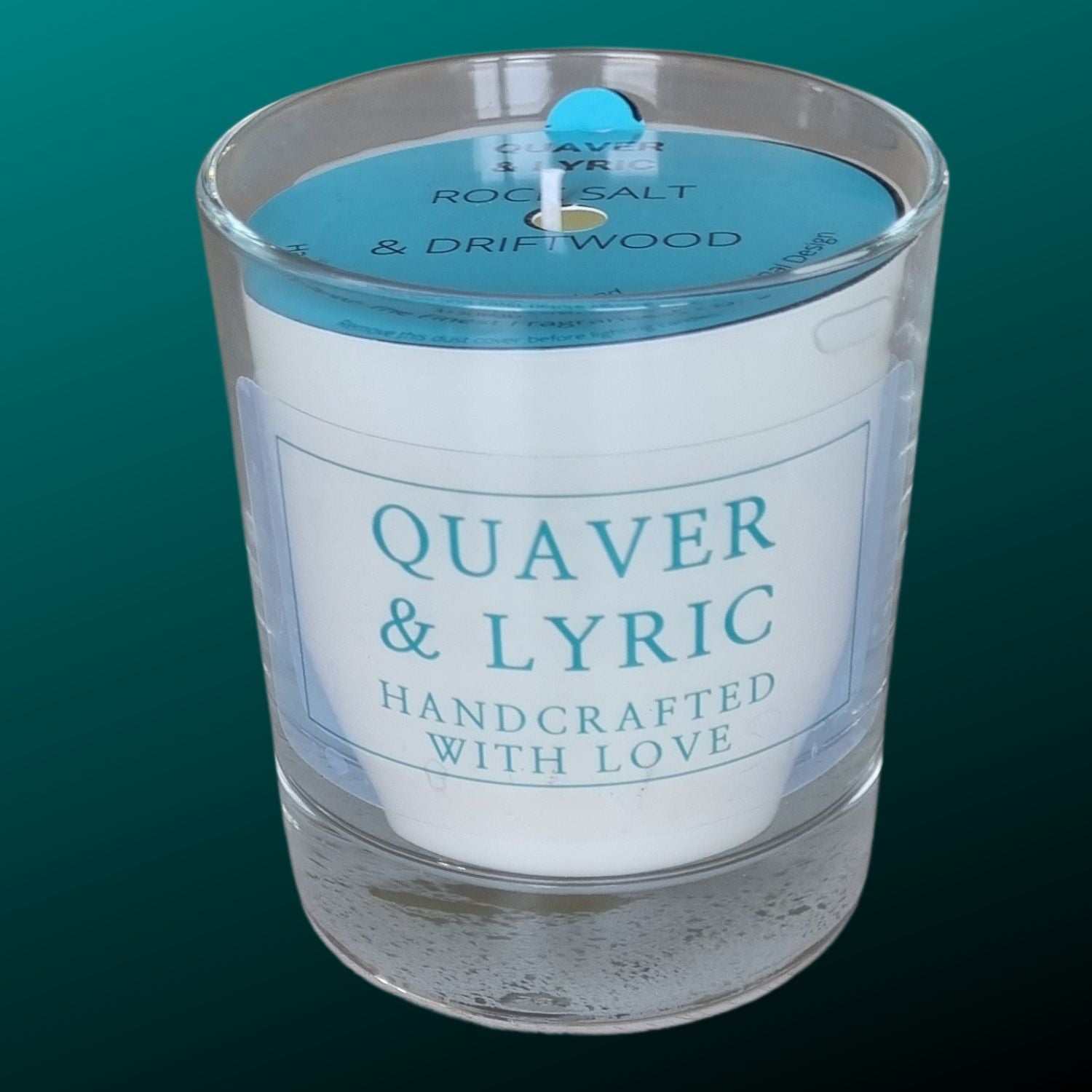 a white scented candle in a glass container with Quaver&Lyric Handcrafted with love label in teal on a transparent background. With a blue dust cover