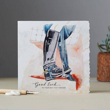 A greeting card with a watercolor illustration of legs in equestrian riding boots. Text on the card reads Good luck—put your best foot forward. A plant and pencils are nearby.