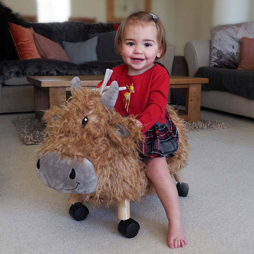 A smiling toddler wearing a red top and plaid skirt sits barefoot on a fuzzy toy bull with wheels, indoors in a cozy living room with sofas and cushions in the background.