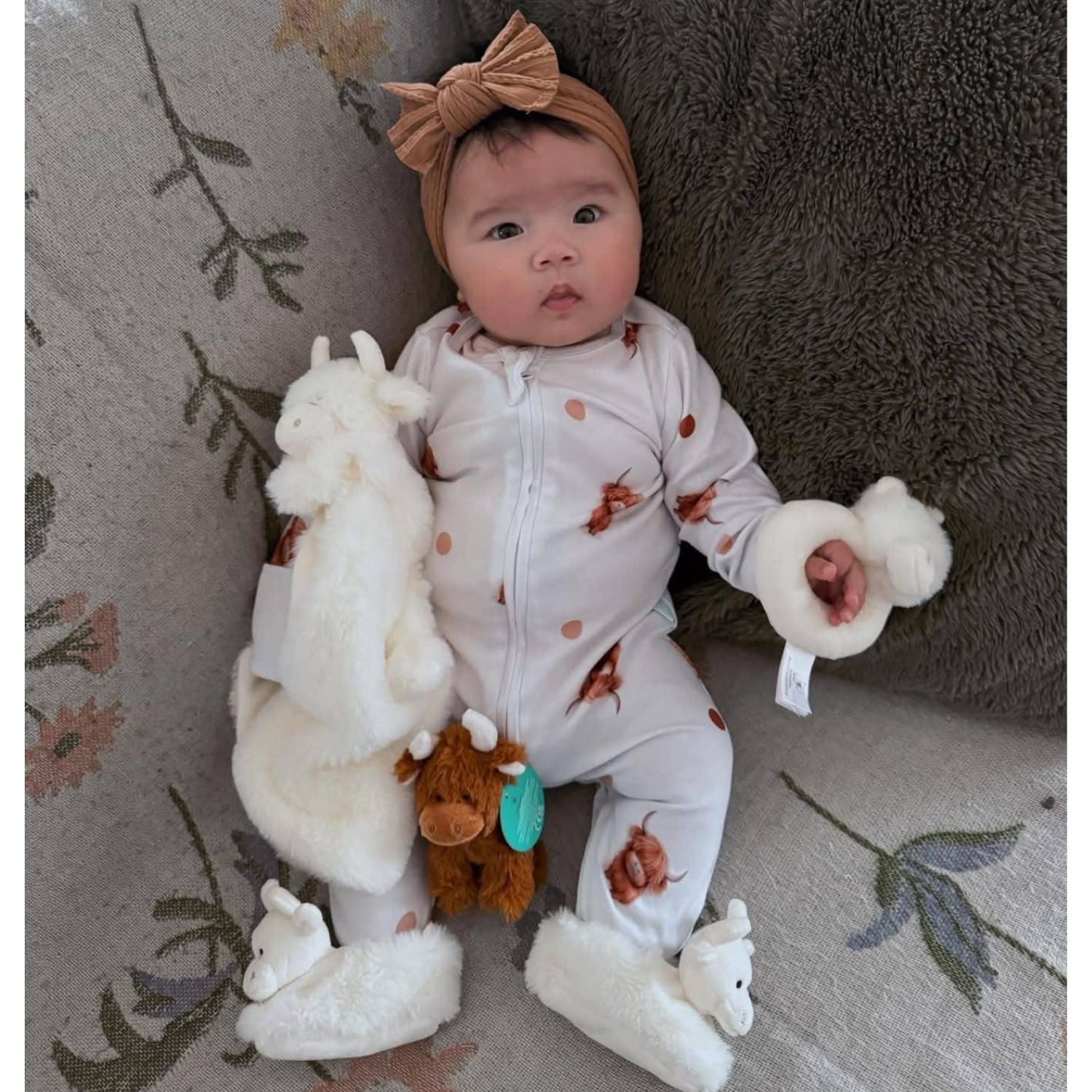 a baby with a collection of newborn toys including cream highland cow rattle, comforter toy with blanket and slippers