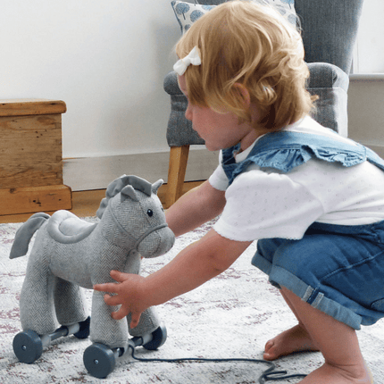 A young child with blonde hair and a white bow plays with a gray plush horse toy on wheels while kneeling on a carpeted floor in a cozy living room.