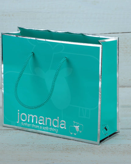 A turquoise gift bag with silver edges and teal rope handles, featuring jomanda, the slogan Softer than a soft thing, and a sheep designâ€”ideal for gifting the Highland Cow Toy Gift Set by Jomanda Soft Toys & Accessories.