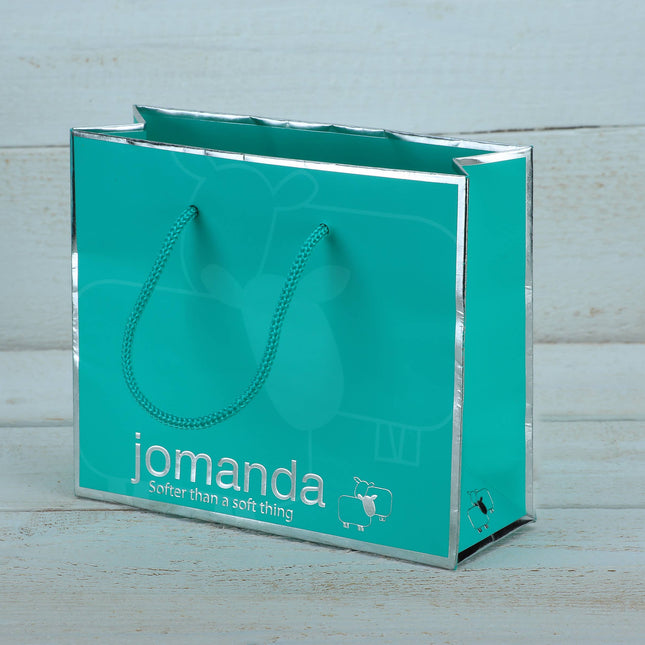 A turquoise gift bag with silver edges and teal rope handles, featuring jomanda, the slogan Softer than a soft thing, and a sheep designâ€”ideal for gifting the Highland Cow Toy Gift Set by Jomanda Soft Toys & Accessories.