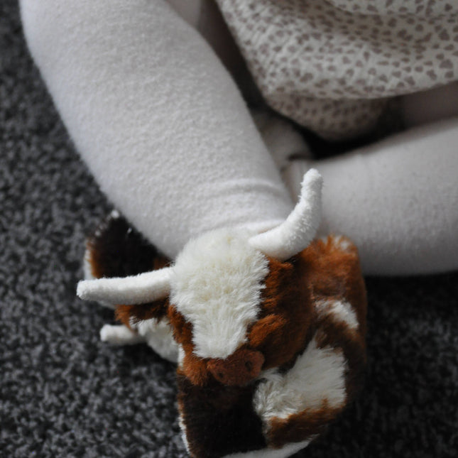 A child in white tights and a patterned dress sits on a gray carpet, wearing Jomanda Soft Toys & Accessories Texas Longhorn Cow Slippers For Infants.