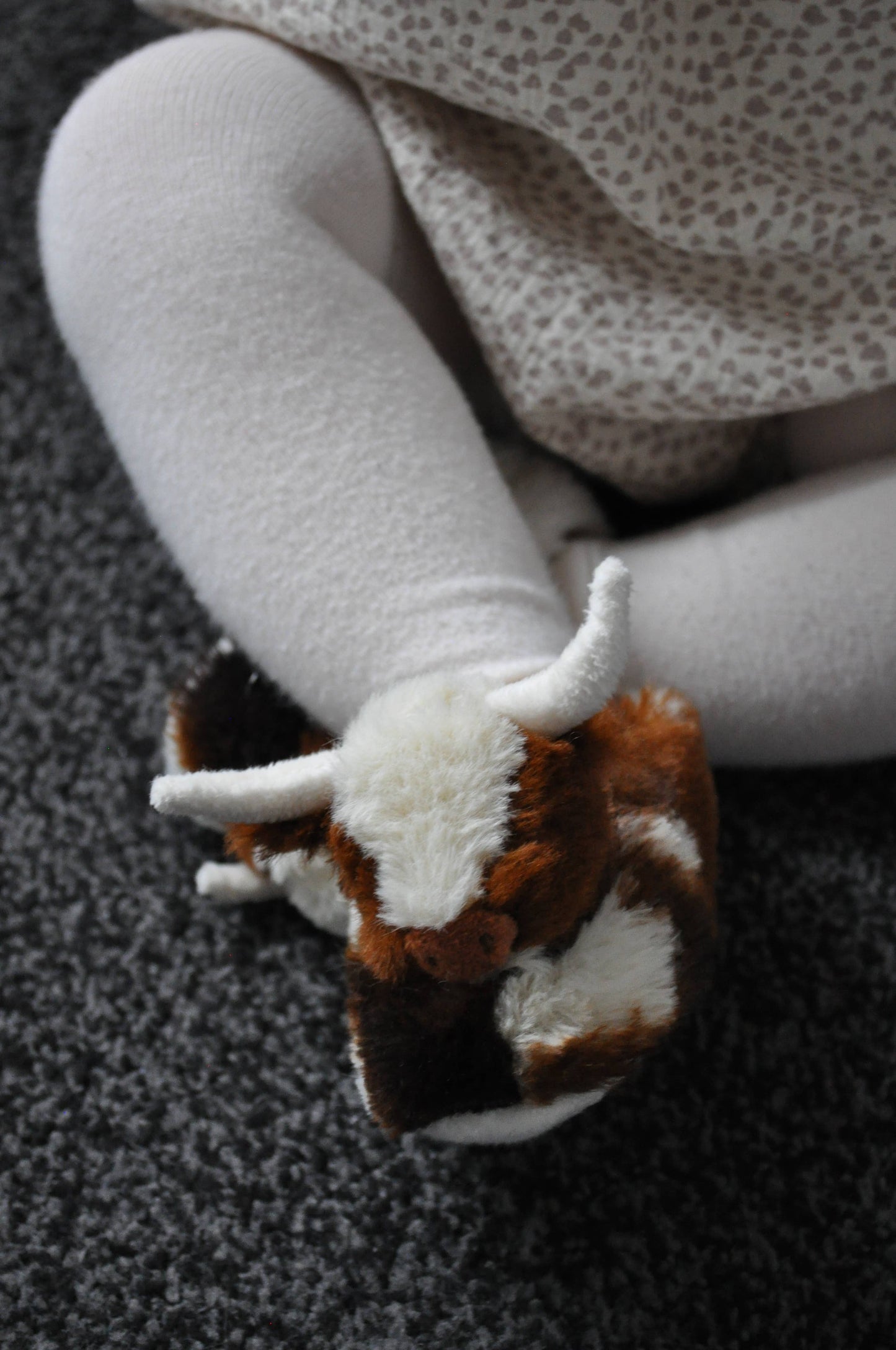 A child in white tights and a patterned dress sits on a gray carpet, wearing Jomanda Soft Toys & Accessories Texas Longhorn Cow Slippers For Infants.