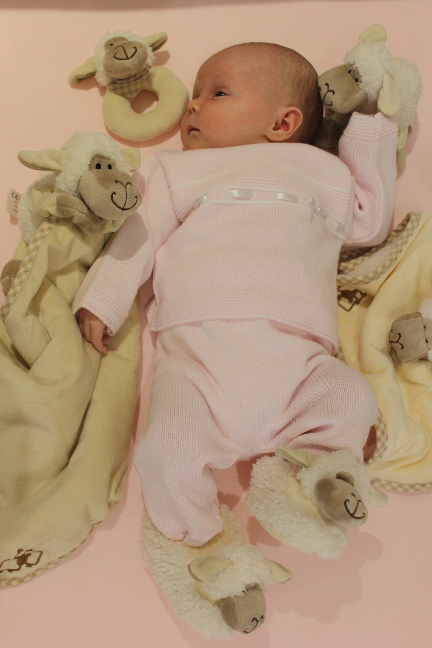 a baby surrounded by cream sheep toys including a rattle and a toy and blanket combo wearing sheep slippers
