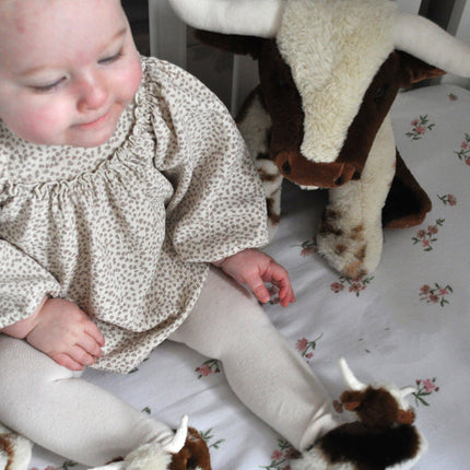 A baby in a crib wears Texas Longhorn Cow Slippers For Infants by Jomanda Soft Toys & Accessories, beside a stuffed cow on white floral beddingâ€”a charming scene ideal for an infant gift.
