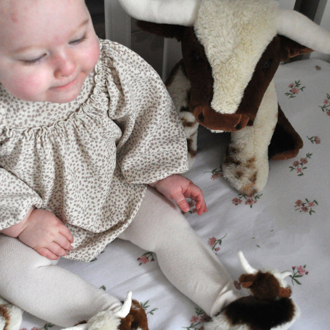 A baby in a crib wears Texas Longhorn Cow Slippers For Infants by Jomanda Soft Toys & Accessories, beside a stuffed cow on white floral beddingâ€”a charming scene ideal for an infant gift.