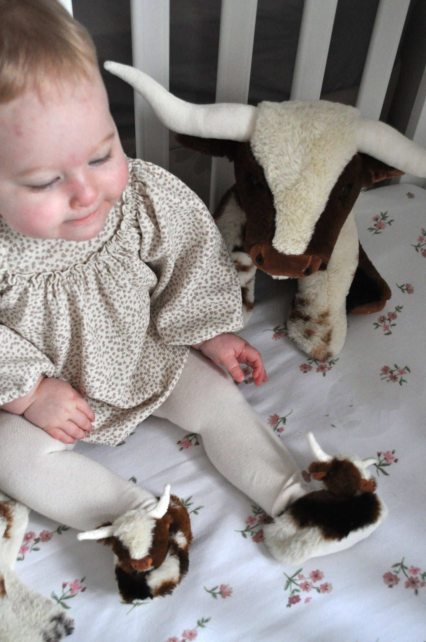 A baby in a crib wears Texas Longhorn Cow Slippers For Infants by Jomanda Soft Toys & Accessories, beside a stuffed cow on white floral bedding—a charming scene ideal for an infant gift.