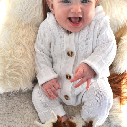 A smiling baby in a white knit onesie with brown buttons wears Jomanda Soft Toys & Accessories Texas Longhorn Cow Slippers for Infants while sitting on a soft rugâ€”an adorable and ideal gift.