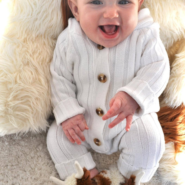 A smiling baby in a white knit onesie with brown buttons wears Jomanda Soft Toys & Accessories Texas Longhorn Cow Slippers for Infants while sitting on a soft rugâ€”an adorable and ideal gift.