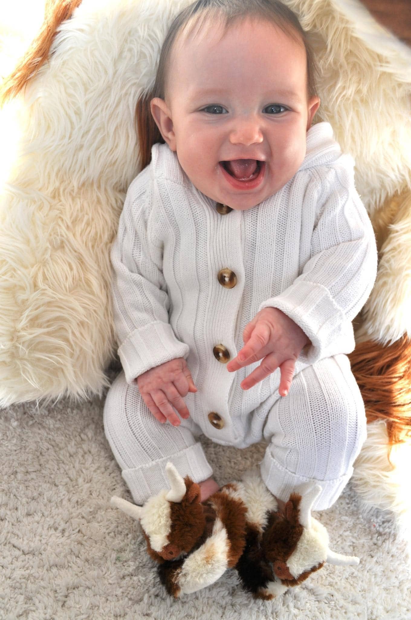 A smiling baby in a white knit onesie with brown buttons wears Jomanda Soft Toys & Accessories Texas Longhorn Cow Slippers for Infants while sitting on a soft rug—an adorable and ideal gift.