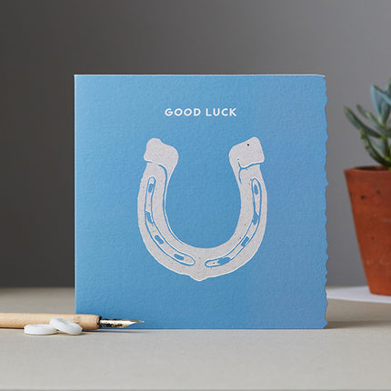 The Equestrian Greeting Card Good Luck Horseshoe Blank by Deckled Edge Ltd features a white horseshoe and GOOD LUCK on a blue background—perfect for well wishes. Inspired by Janna Cossettini, it’s shown next to a pen and potted succulent.