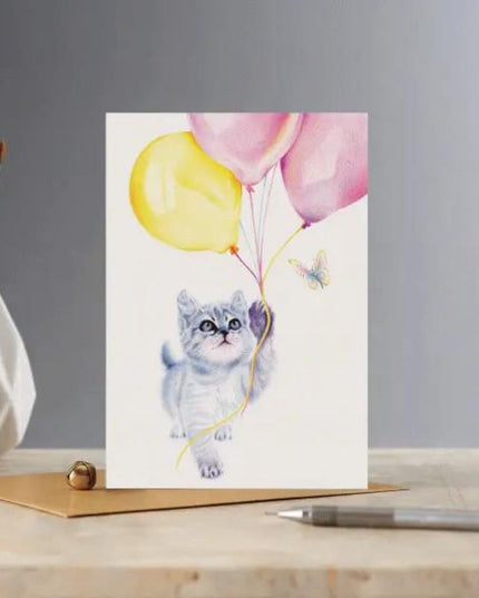 The Cat Greeting Card Kitten and Balloons Blank Deckled Edge by Deckled Edge Ltd features a Janna Cossettini grey tabby kitten with balloons and a butterfly, set on a desk with pen, envelope, and paintbrush holder in the background.
