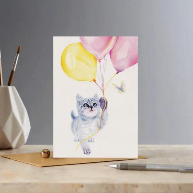 The Cat Greeting Card Kitten and Balloons Blank Deckled Edge by Deckled Edge Ltd features a Janna Cossettini grey tabby kitten with balloons and a butterfly, set on a desk with pen, envelope, and paintbrush holder in the background.