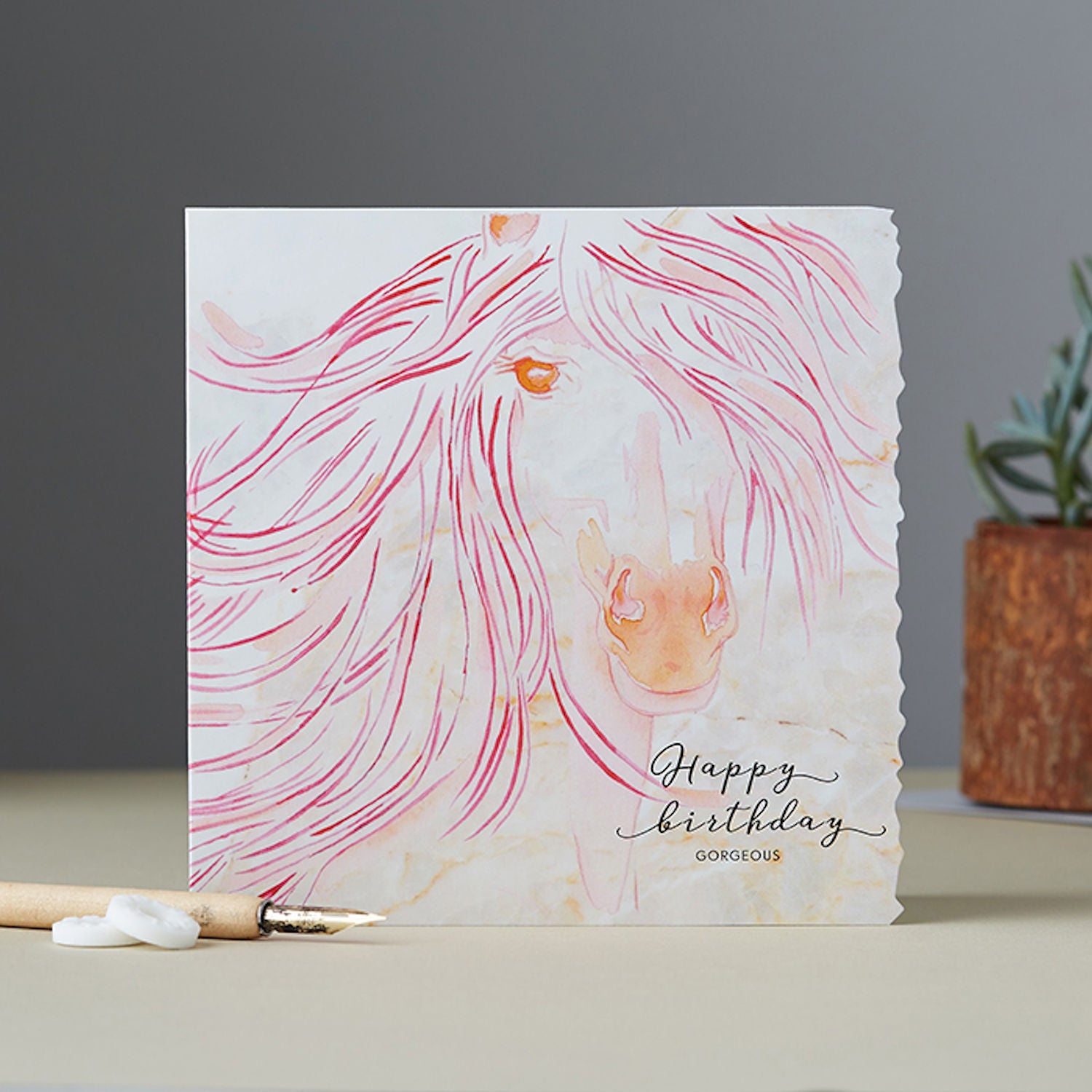 a square greeting card with a crinkled edge on one side resting on a surface with an ink pen and polos in front and a planr behind. The card features a beautiful horse with a flowing mane in pink and orange with wording on the outside Happy Birthday Gorgeous