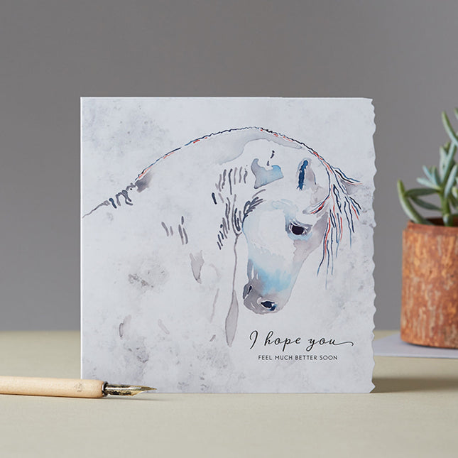 The Horse Get Well Greeting Card Deckled Edge by Deckled Edge Ltd features a gentle watercolor white horse illustration by Janna Cossettini and the message I hope you feel much better soon. Pen and potted plant shown for display.