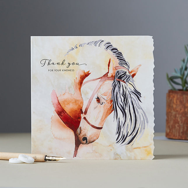 The Greeting Card Blank Inside Deckled Edge Horse Thank You by Deckled Edge Ltd features a watercolor bay horse and Thank you for your kindness message, with a pencil, two white stones, and a blurred potted plant in the background.