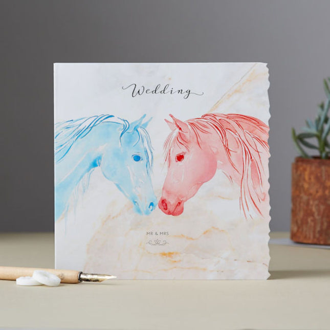 The Equestrian Wedding Greeting Card Deckled Edge by Deckled Edge Ltd features Janna Cossettini’s art—two watercolor horses, blue and pink, touching noses beneath a cursive Wedding. Perfect for wedding wishes.