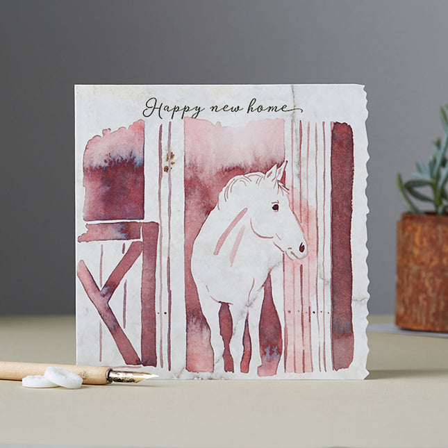 The Horse Greeting Card Happy New Home by Deckled Edge Ltd features a watercolor white horse in a barn doorway with “Happy New Home” handwritten at the top, complete with charming pencil and plant accents.
