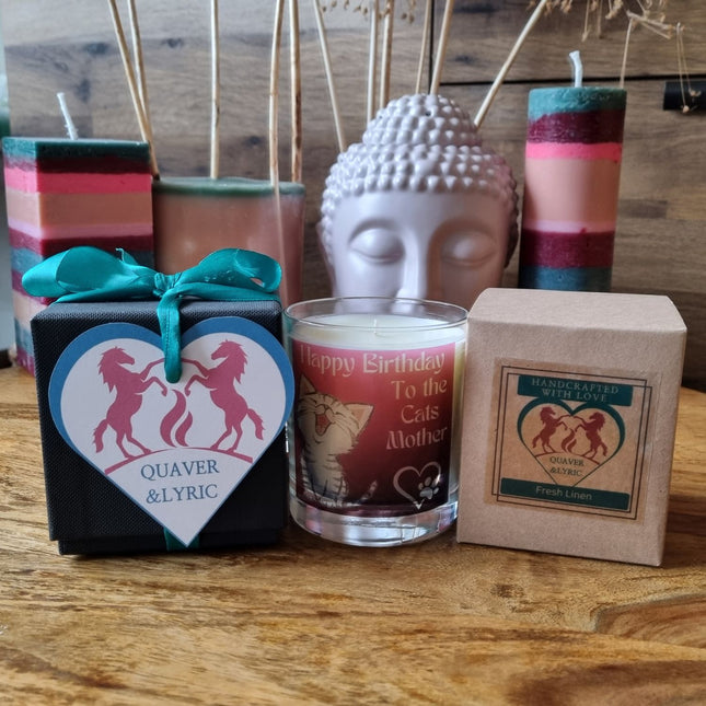 Three candles are shown: a handmade one with a heart label, Quaver&Lyric’s Scented Candle In Glass Container Happy Birthday Cats Mother Gift for cat mums, and a Fresh Linen boxed candle. A Buddha head and colored candles appear in the background.