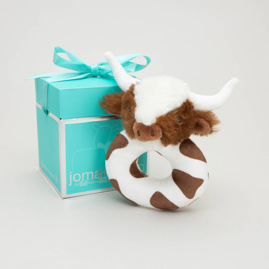 a soft plush circular brown and white baby rattle with a fluffy brown and white longhorn cow head next to a jomanda branded gift box