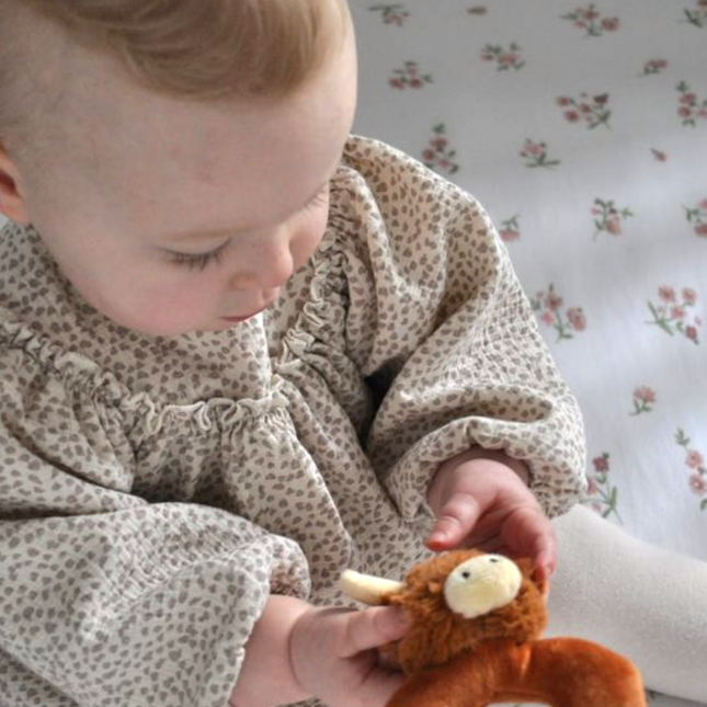 a baby playing with a donut shaped new baby rattle in brown soft plush fabric with a cute brown highland cows head with cream horns and ears