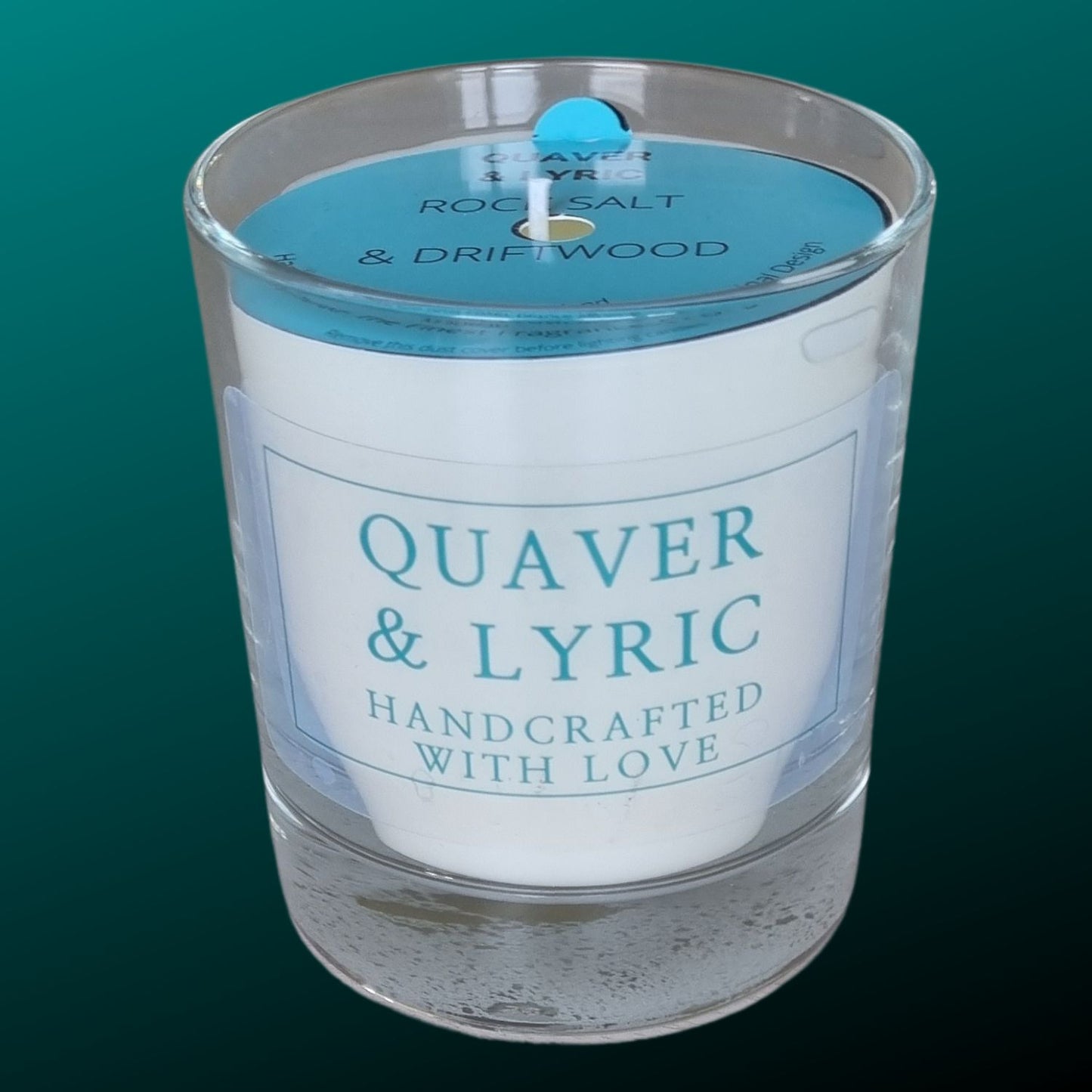 a candle in a glass with quaver & lyric handcrafted with love label and blue dust cover on a teal background
