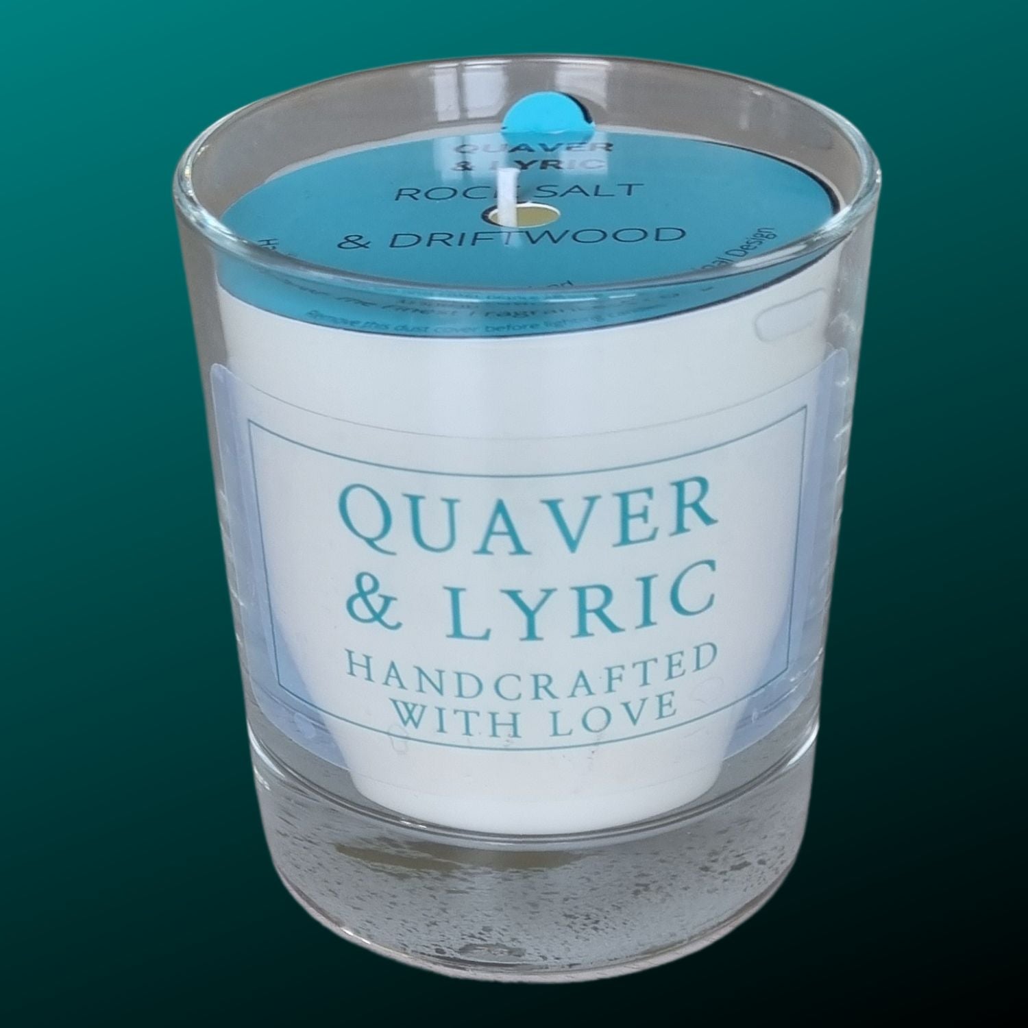 a candle in a glass with quaver & lyric handcrafted with love label and blue dust cover on a teal background