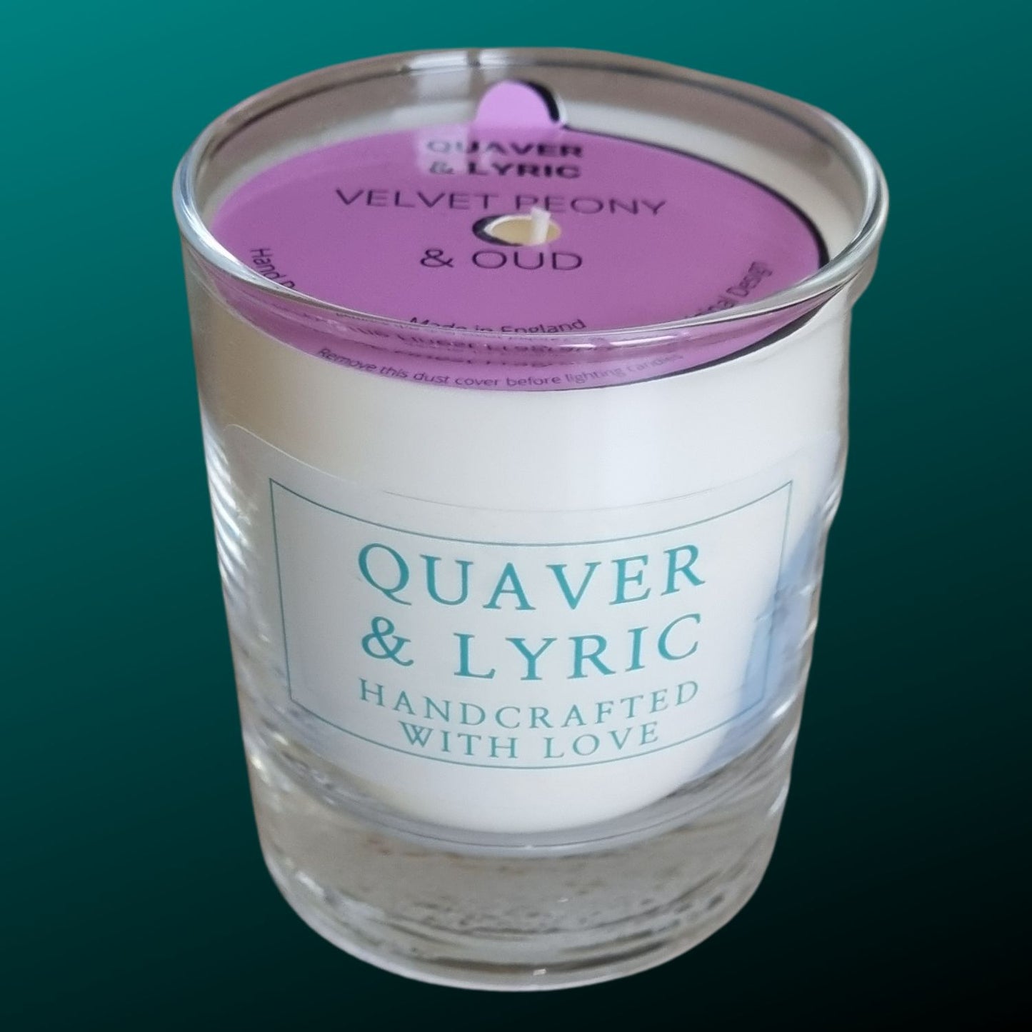 a candle in a glass with quaver & lyric handcrafted with love label and pink dust cover on a teal background