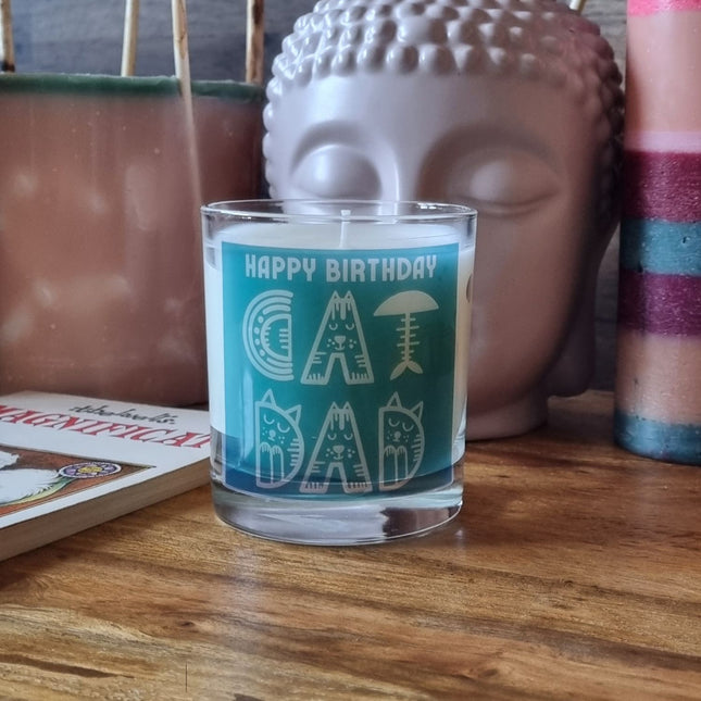 The Quaver&Lyric Scented Candle In Glass Container, featuring a teal Happy Birthday Cat Dad label, makes the perfect birthday gift and is displayed on a wooden surface beside decor items and a greeting card.