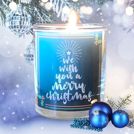 The Quaver&Lyric Christmas Scented Candle Wish You A Merry Christmas Glass Container Gift is displayed among blue ornaments, beads, frosted pine branches, and festive décor, with a sparkling tree in the background.