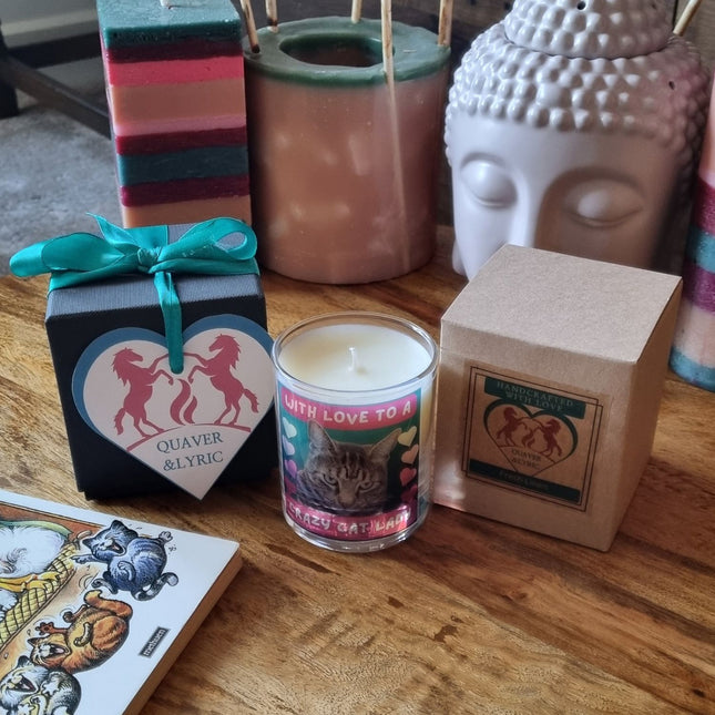 A Quaver&Lyric Scented Candle In Glass Containerâ€”Crazy Cat Lady Bengal Cat Designâ€”sits between a black gift box with a unicorn heart label and a brown box, among decorative candles and premium fragrances on a wooden table.