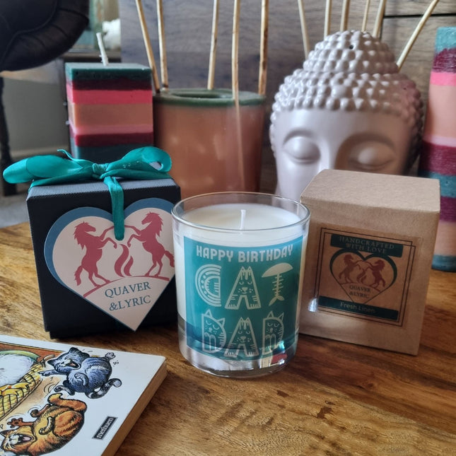 A Quaver&Lyric Scented Candle In Glass Container—Happy Birthday Cat Dad Gift—is displayed with a heart-shaped box, wildflower soap, and stacked handmade candles beside a Buddha head sculpture on a wooden table.