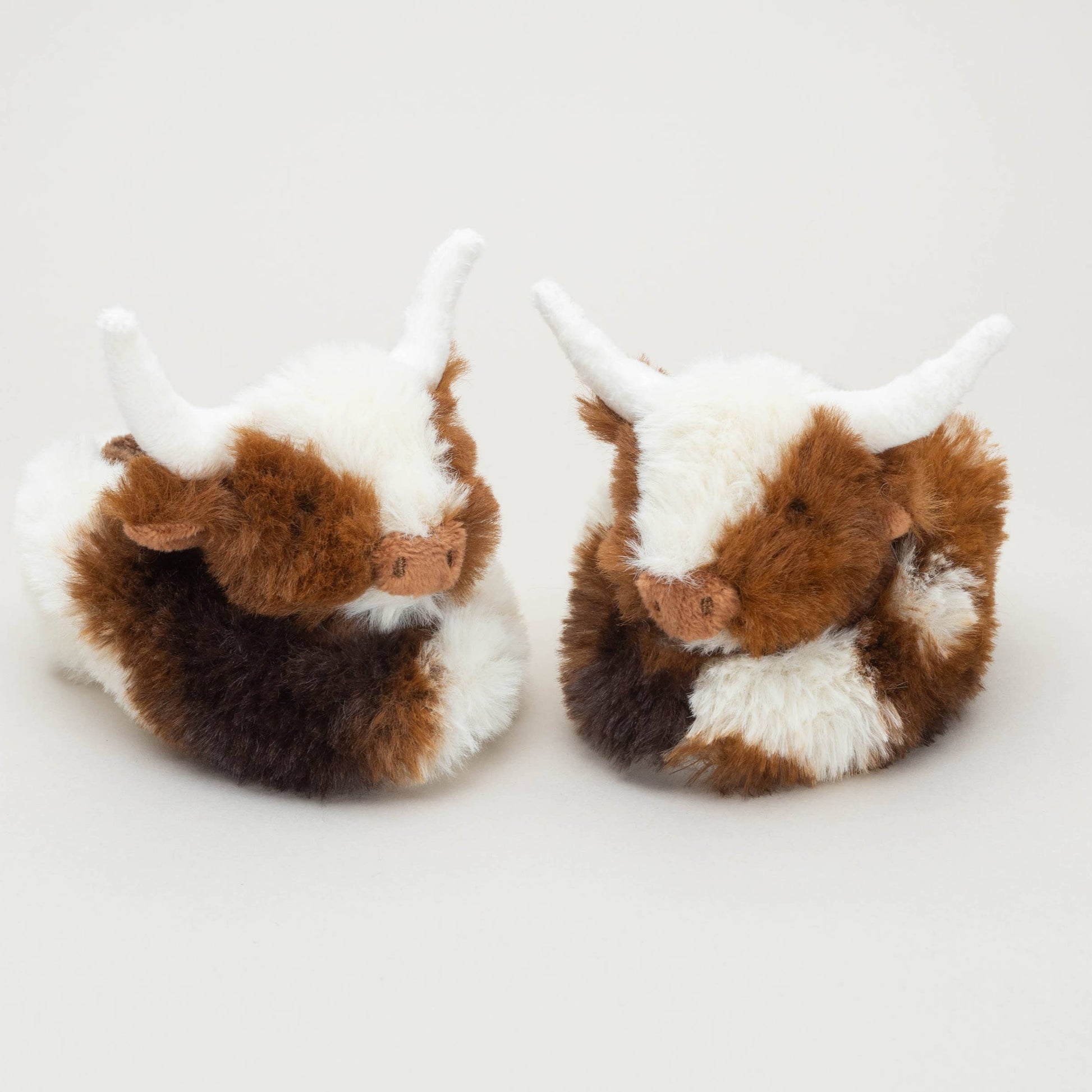Two Texas Longhorn Cow Slippers For Infants by Jomanda Soft Toys & Accessories, featuring cute brown and white fur with white horns, are placed side by side on a plain background—an ideal gift or cozy baby booties.