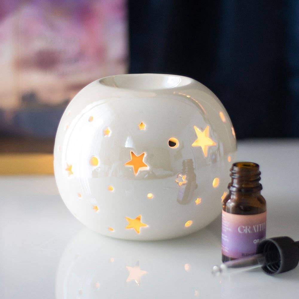 a white spherical wax warmer with stars cut out and and irridescent finish next to a bottle of fragrance oil