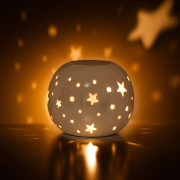 a white spherical wax warmer with stars cut out and and irridescent finish being used in the dark showing the projections of stars
