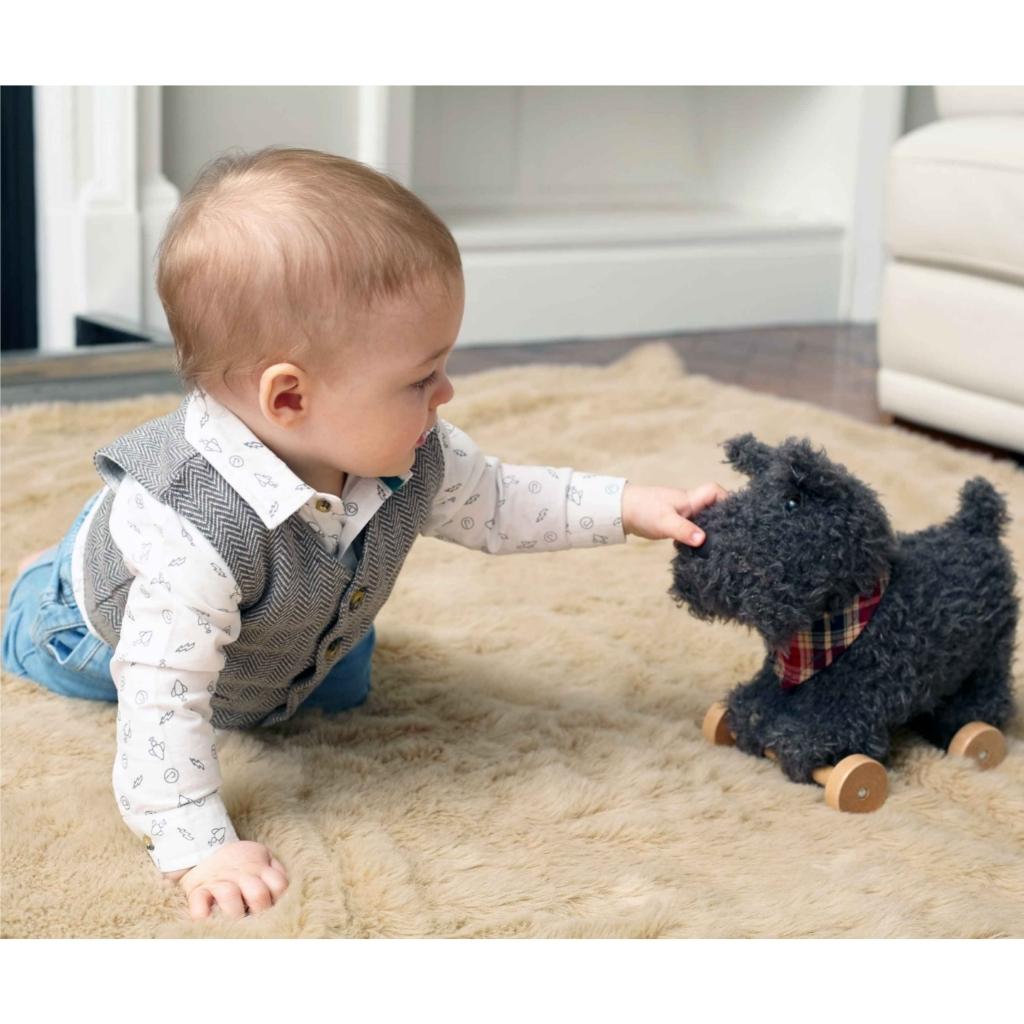 a baby boy playing with a dark grey scottie dog soft toy on wheels
