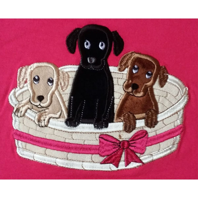 Three embroidered puppiesâ€”a beige, a black, and a brownâ€”sit in a white basket with a pink ribbon on a pink background. This Dog T Shirt by British Country Collection is perfect for any young dog lover.
