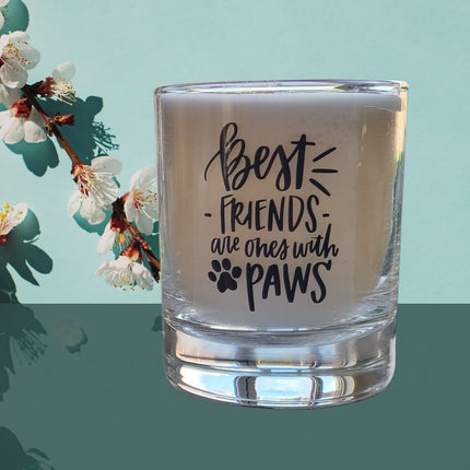 A Quaver&Lyric Scented Candle In Glass Container, featuring a â€œBest friends have pawsâ€ pet design, sits on a reflective surface with white cherry blossoms and a teal blue background. The creamy candle has a high quality wax blend.