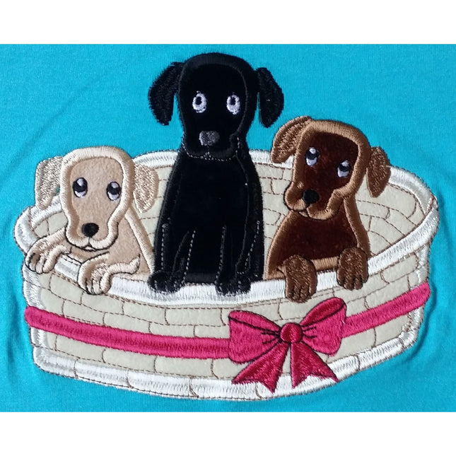 This British Country Collection Dog T Shirt for children features three embroidered puppiesâ€”a beige, black, and brownâ€”sitting in a cream basket with a pink ribbon and bow, set against a blue background.