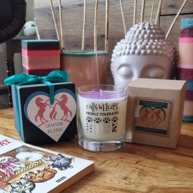 A Quaver&Lyric Funny Cat Lover Gift Scented Glass Candle Cats Welcome 30cl, a heart-shaped horse box, a brown candle, colorful candles, a Buddha head, incense sticks, and a bright card decorate the wooden table.