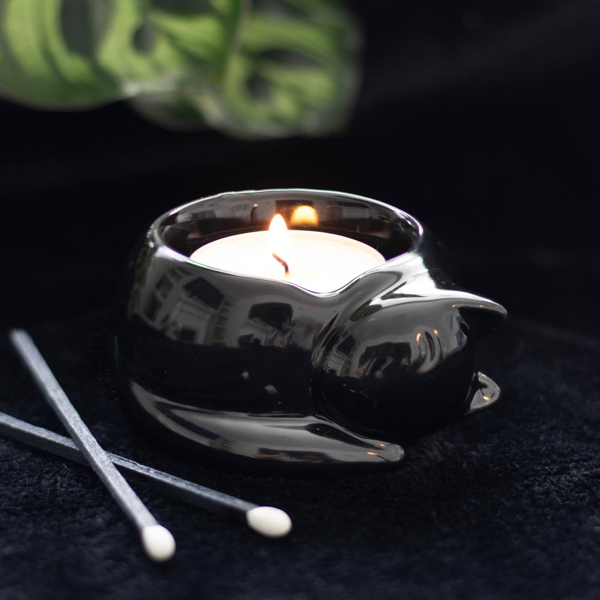 a black glossy tealight holder in the form of a curled up cat with a lit tealight inside and two long matches lying by the side