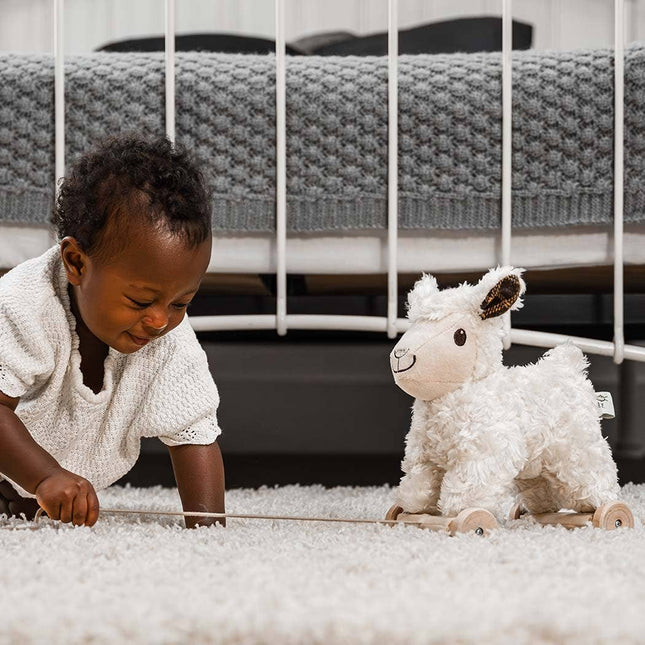 A baby in white smiles while pulling the Little Bird Told Me Uk Pull Along Toy Sheep Lambert, enjoying tactile playtime in a cozy nursery, with a crib in the background.