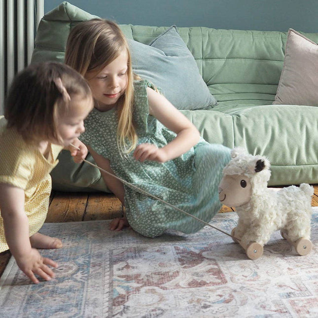 Two young children play on a rug, pulling the Pull Along Toy Sheep Lambert by Little Bird Told Me UK. The fuzzy white sheep has wheels and a string, with a light green couch and pastel cushions in the background.