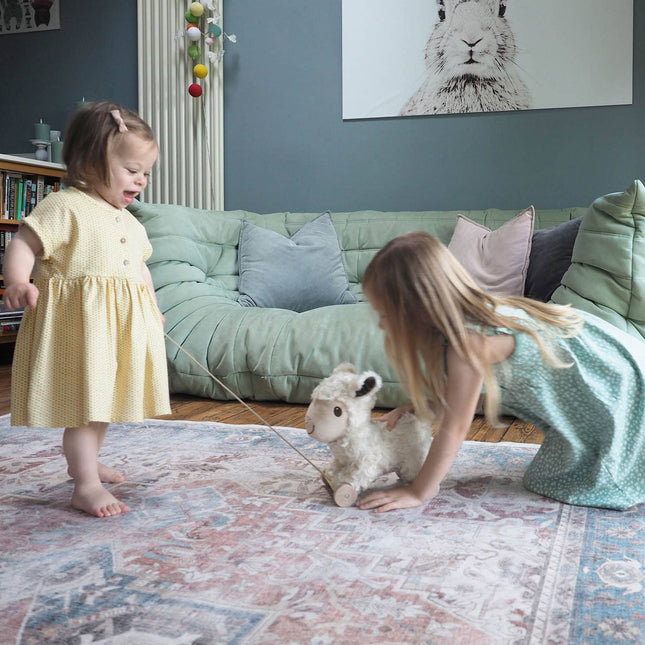 Two young girls play on a patterned rug; one stands in a yellow dress, the other in green kneels and pulls along the Little Bird Told Me UK Pull Along Toy Sheep—ideal for toddler development. A cozy sofa and rabbit picture are in the background.