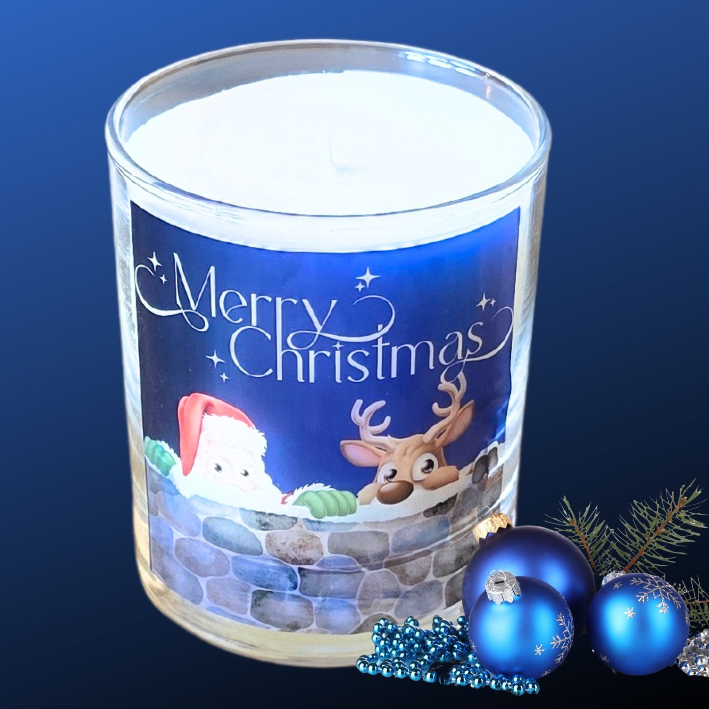 A Christmas scented candles with a charming Merry Christmas design featuring Father Christmas and a reindeer peeping over a wall. The candle is next to festive blue baubles.