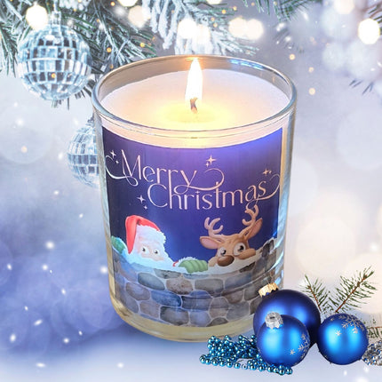 A lit Christmas scented candles with a charming Merry Christmas design featuring Father Christmas and a reindeer peeping over a wall. The candle is set in a snowy festive setting.